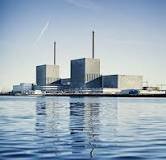 Kärnkraftverk vid vattenkanten, blå himmel, reflektion i vattnet, lugnt väder, industribyggnad med skorstenar.