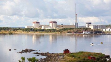 Kärnkraftverk vid kust, röda stugor, stilla vatten, molnig himmel, grönska, reflektioner på vattenytan.