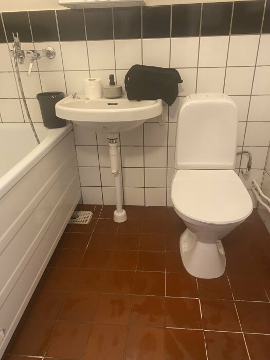 Ett badrum med toalett, handfat, kakelväggar, brunt golv, och en svart väska.