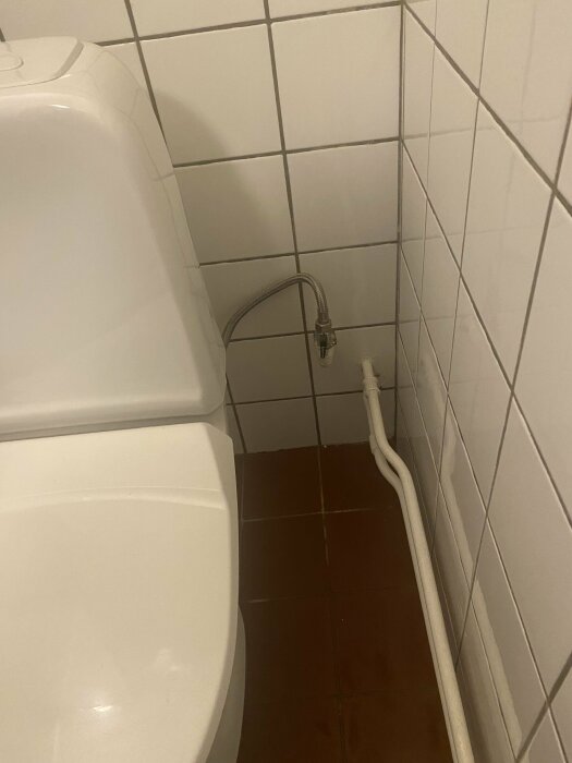 Ett toalettrum med vitt kakel, toalettstol och synliga rör vid sidan.
