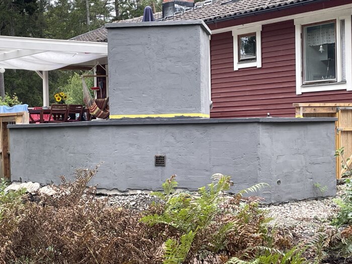 Röd trävilla, grå betongvägg, fönster, veranda med hängmatta, grönska, grus, del av bostadsområde.