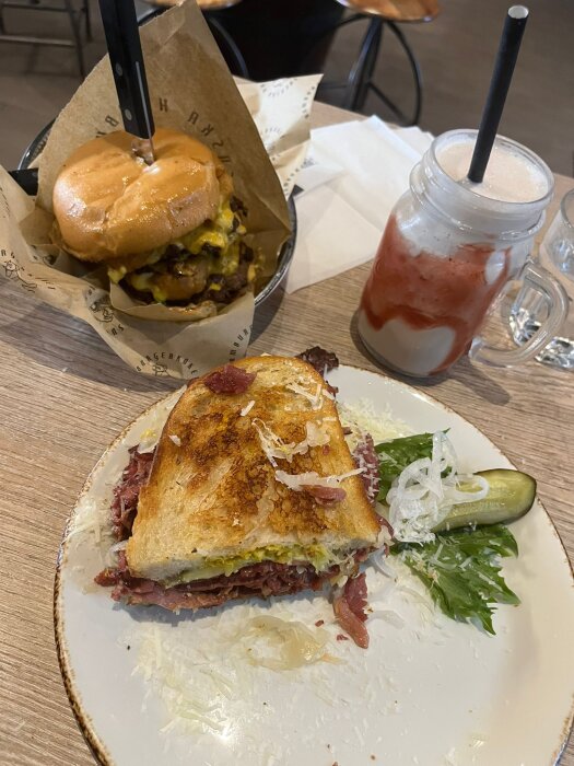 En halverad Reuben-smörgås, hamburgare, pommes frites, röd dryck. Restaurangmiljö. Snabbmat. Sallad och pickles som tillbehör.
