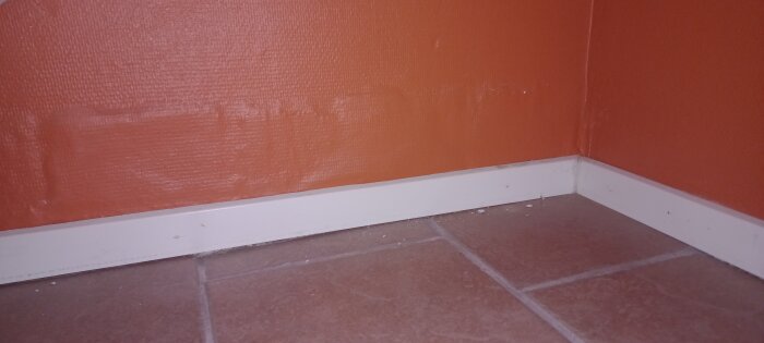 Hörn av rum med orange vägg, vit list, brun klinkergolv.