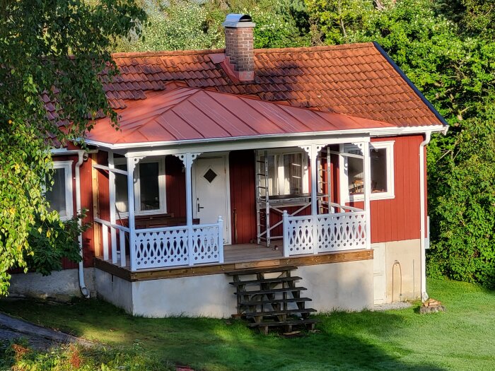 Rött trähus med veranda, vita staket, grönt gräs och tegeltak i solsken.