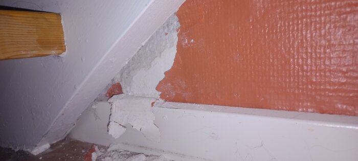 Vägg med avskalad färg och puts, exponerat tegel, vit list, synlig träbit. Behov av reparation eller renovering.