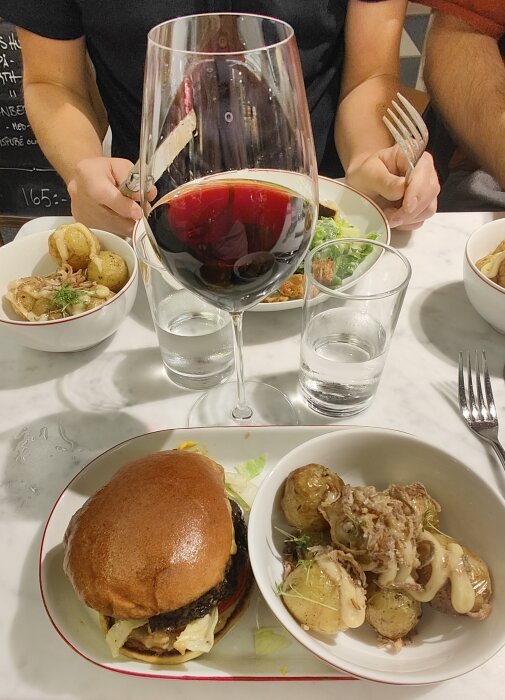 Måltid med hamburgare, sallad, potatisskivor, rödvin och vattenglas på ett marmorbord, två personer som äter.