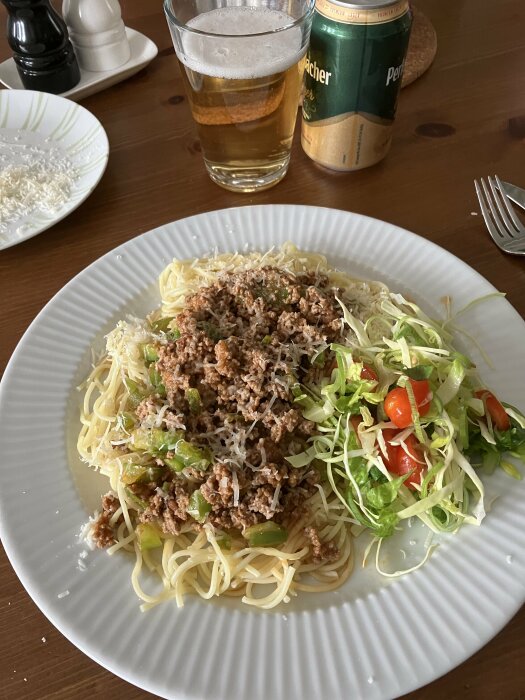 Spaghetti med köttfärssås, riven ost, sallad, öl och bordskryddor i bakgrunden.