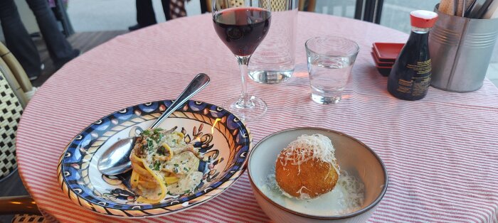 Ett restaurangbord med pasta, arancini, rödvin, vatten och sojasås på rödvit rutig duk.