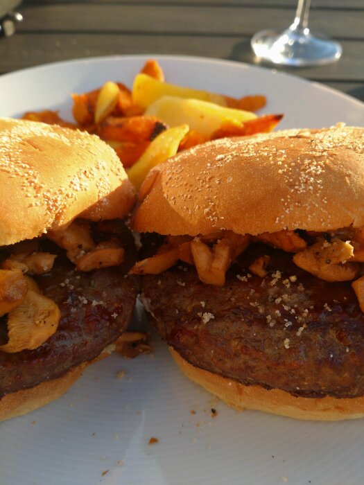 Två hamburgare med stekta svampar och pommes frites på en tallrik i solljus, glas i bakgrunden.