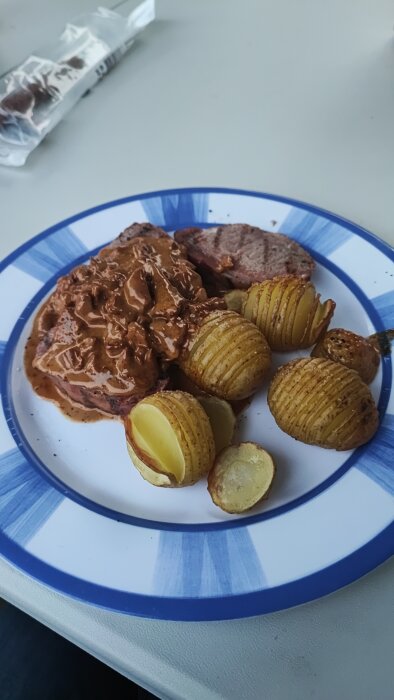 En tallrik med hasselbackspotatis och kött i gräddsås på en vit bordyta.