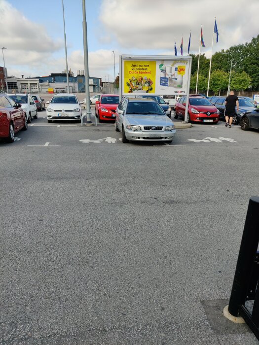 Bilparkering, felaktigt parkerad bil, reklamskylt, byggnader i bakgrunden, person, dagtid, delvis molnigt, asfalt.