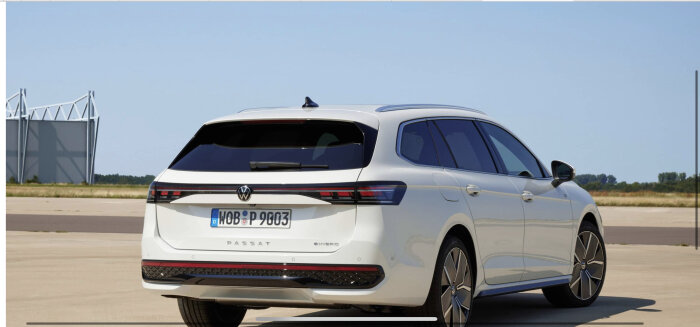 En vit Volkswagen Passat kombi, hybrid, parkerad utomhus med klart himmel och industribyggnad i bakgrunden.