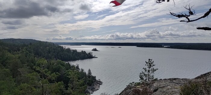 Utsikt över havet med öar, klippor och skog, delvis molnigt, svenska flaggan fladdrar i förgrunden.