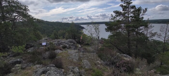 Skogbevuxen klippa, sjöutsikt, moln, informationsskylt, flagga, vild natur, dagtid, orört landskap.