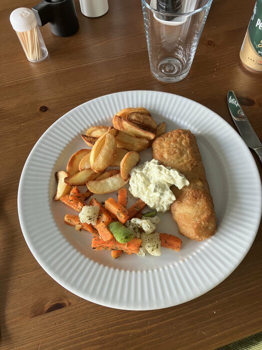 Tallrik med mat: stekt fisk, potatis, morötter, remouladsås. Glas vatten och kryddburkar i bakgrunden.