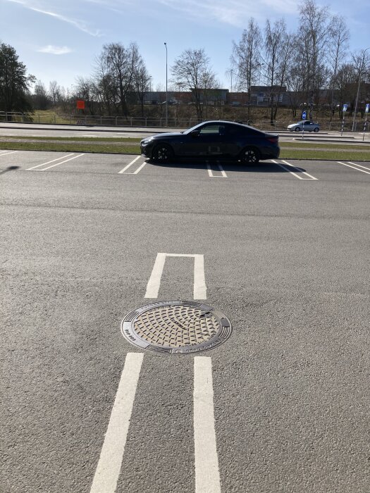 Asfalterad väg, parkerad bil, vägmarkeringar, brunnlock, träd, klar himmel, dagtid.