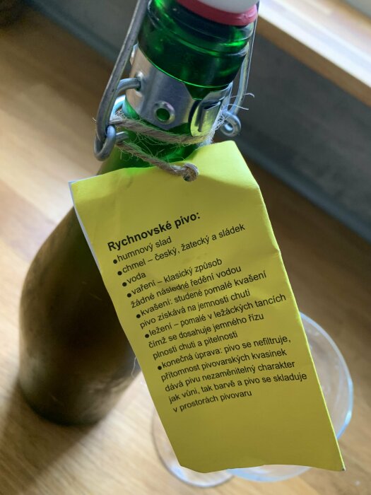 Grön ölflaska med etikett, metallklämma, text på tjeckiska, beskriver öltillverkningsprocess.