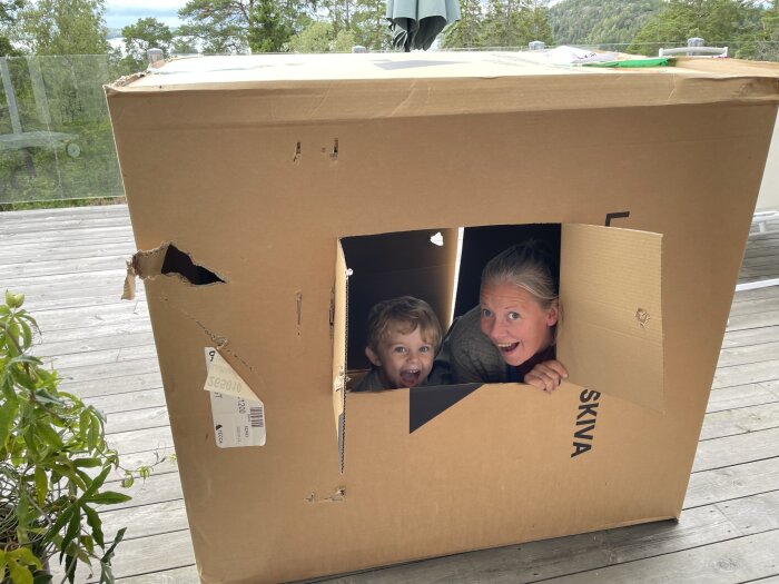 Två personer skrattar inne i en stor kartongkasse utomhus på en terrass.