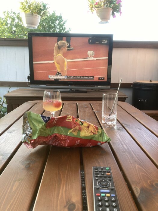 Utomhusmiljö, TV med tennis, snacks, fjärrkontroll, tomma glas, träbord, avslappnad stämning.