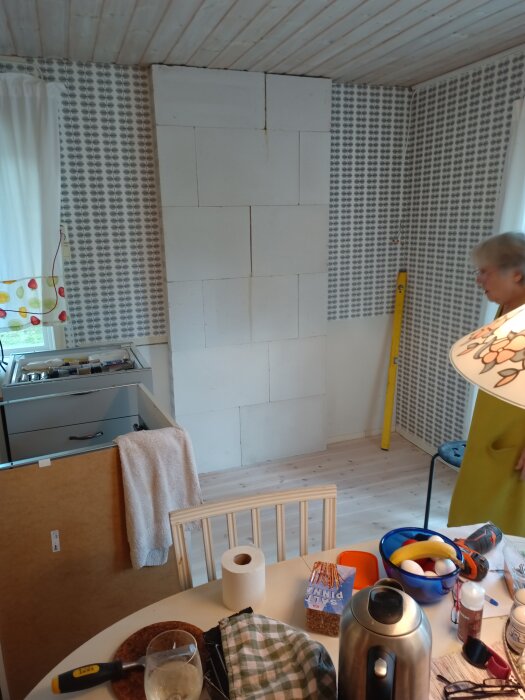 Ett rum under renovering med oavslutad vägg, person delvis synlig, byggnadsmaterial och hushållsartiklar framme.