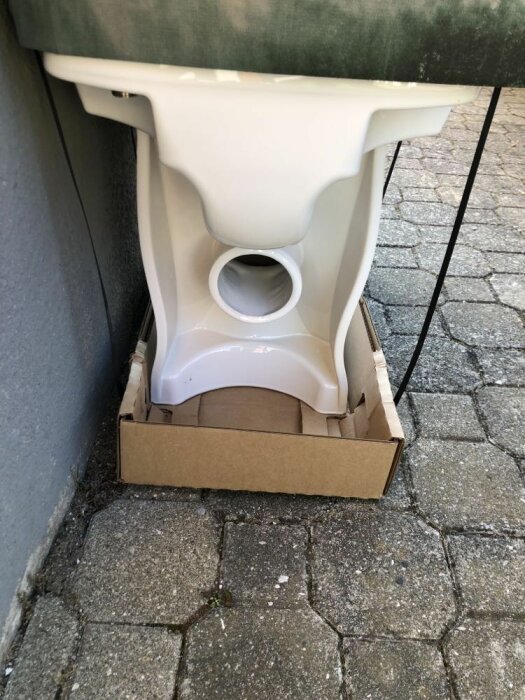 En toalettstol utan cistern står på en kartong mot en vägg utomhus.