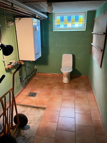 En spartansk toalett i ett källarutrymme med gröna väggar, rörgenomföringar och en dörr i bakgrunden.