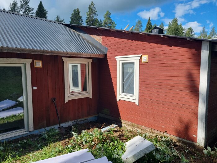 Rödlackerat trähus, plåttak, fönster, skogsbakgrund, soligt, oarbetad mark förgrund, spade lutad mot hus.