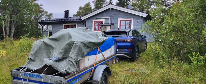 Grått hus med röda fönsterkarmar, släpvagn, bil, natur. Nummer 13 synligt på huset. Overcast väder, orörd natur.