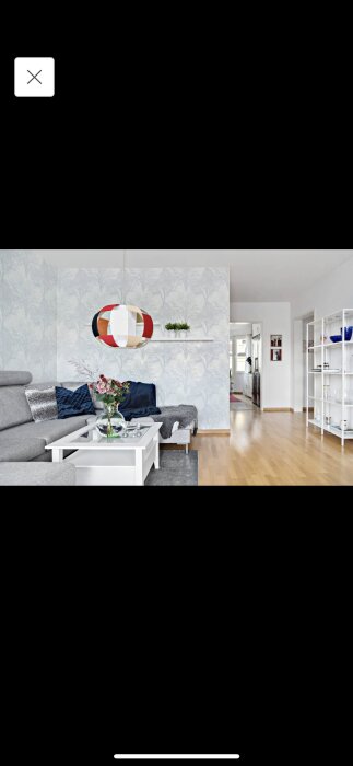 Modernt vardagsrum, stilren design, grå soffa, vit soffbord, trägolv, vägghylla, färgglad taklampa, växttapet, ljus inredning.