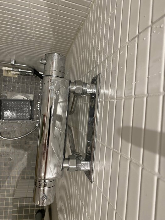 Kromad termostatblandare i en dusch, vit kakelvägg, vattendroppar, och reglaget visar temperaturskala.