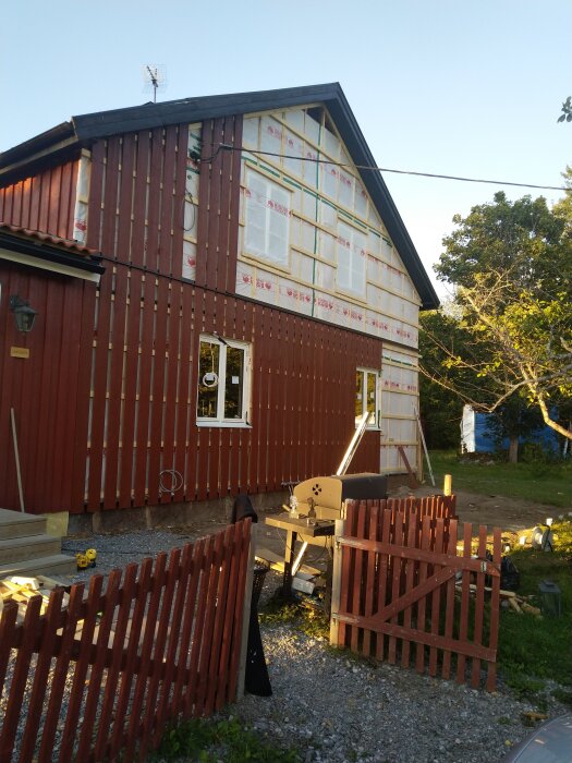 Tvåvåningshus under renovering, rödmålat, vindskydd, trämaterial och verktyg synliga, byggprojekt, trästaket, blå himmel.