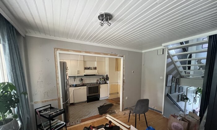 Inomhusrenovering; avskalade väggar, kök, rörigt, stege, byggmaterial, vit takpanel, gardiner, växter, blå soffa.