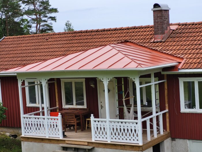 Rött trähus med veranda, vita staket, tak i tegel, skorsten, fönster, natur i bakgrunden.