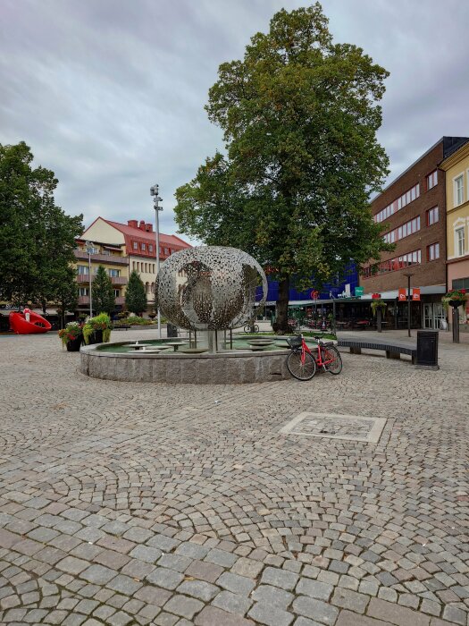 Skulptur på torg, träd, klassisk stenläggning, cykel, byggnader, molnig himmel.