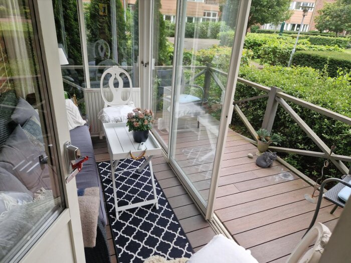 Inredd balkong, trämöbler, vit dyna, blommor, kudde, glaspartier, trädgård, mönstrad matta, avkopplande.