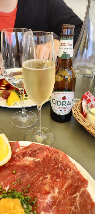 En måltid med skivor av råbiff, citron, ett glas vin, ciderflaska, vattenkaraff och person i bakgrunden.