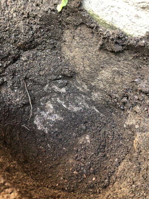 Jordlager och sten nära en betongkant, möjligen en grävd grop eller plantering med synlig markprofil.