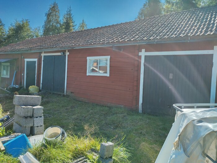 Rödmålat trähus med tegeltak, garageportar, fönster, byggmaterial och grönska i solljus.