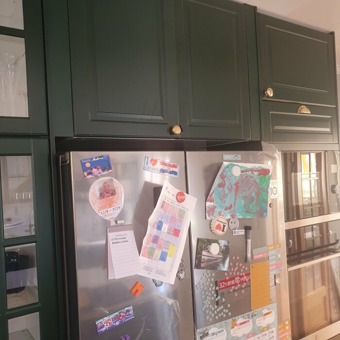 Ett rostfritt kylskåp med magneter och notislappar under gröna köksskåp i ett hem.