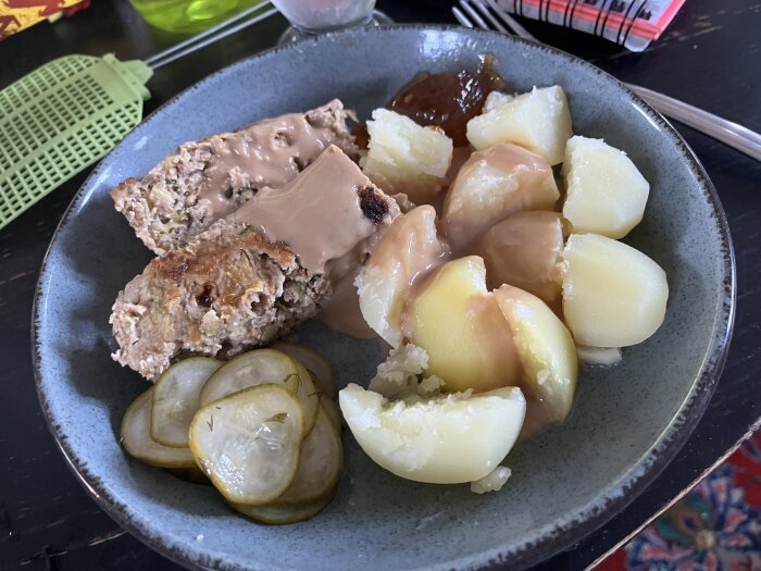 En tallrik med köttfärslimpa, potatis, brun sås och inlagda gurkor på ett bord.