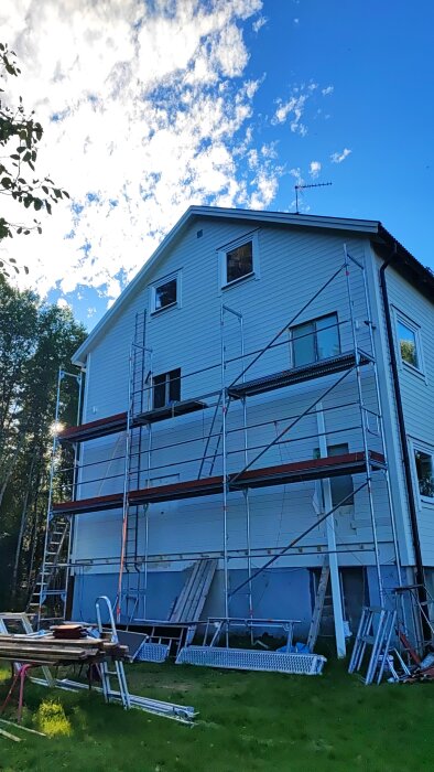 Vitt hus, byggnadsställningar, renovering, grönt gräs, blå himmel med moln, dagtid.