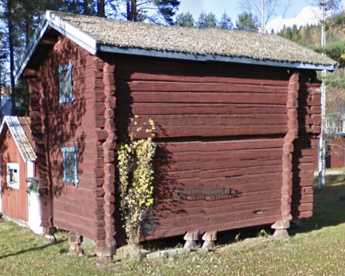 Röd trästuga, torvtak, stenfot, lantlig miljö, solsken, träd, blå himmel, skuggor, traditionell arkitektur.