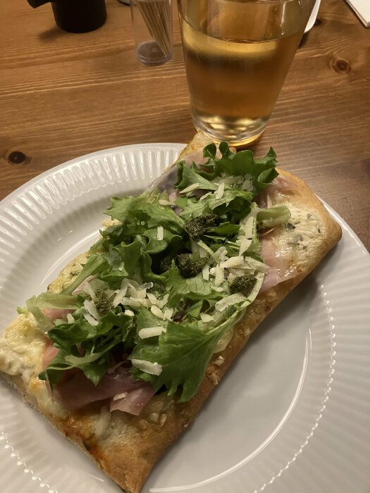Pizza med skinka, ruccola och pesto på vit tallrik, glas med dryck, trämöblerad bakgrund.