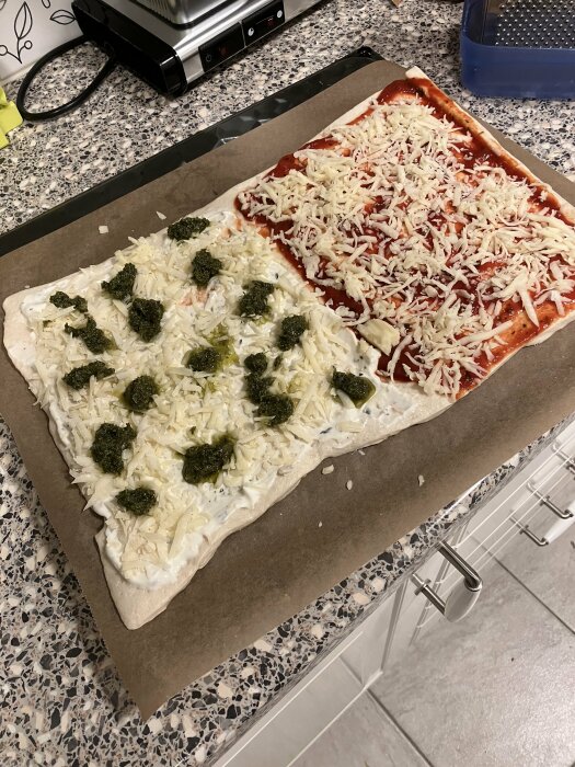 Ogräddad, halvt vitostfylld, halvt röd tomatsåspizza med pestoklickar, på bakplåtspapper, i ett kök.
