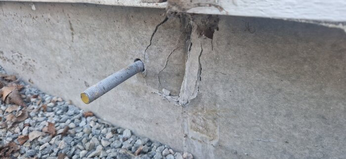 Sprucken betongvägg med utstickande armeringsjärn och grus i förgrunden. Slitage eller skade.
