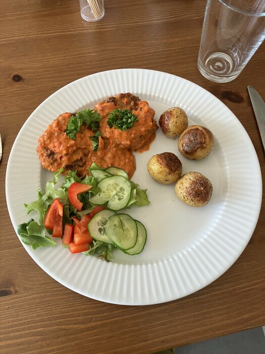 Måltid med köttbullar, röd sås, stekt potatis, sallad, tomat, gurka, persilja, kniv, glas, träbord.