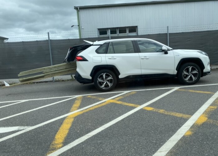 Vit SUV med timmerhalvmeter utsticker från baklucka, parkerad felaktigt över parkeringslinjer, molnig himmel.