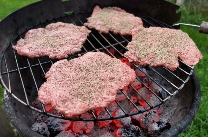 Fyra hamburgare grillas med kryddor på en kolgrill utomhus.