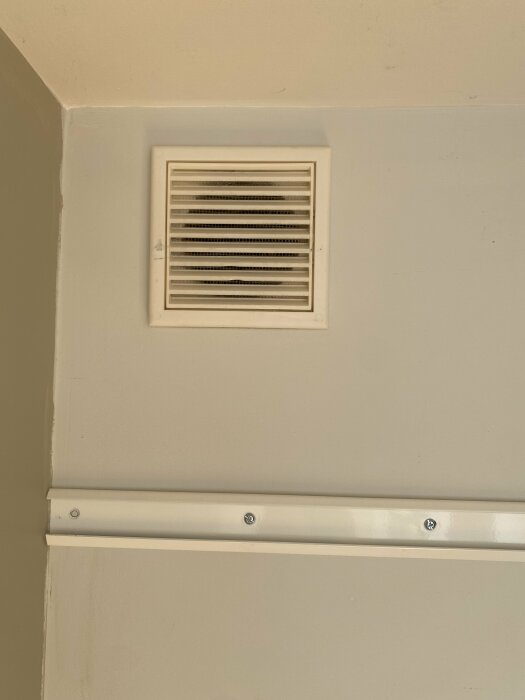 Ventilationsgaller på vägg ovanför vit list, beige vägg, skruvar synliga, inomhus, hörn.