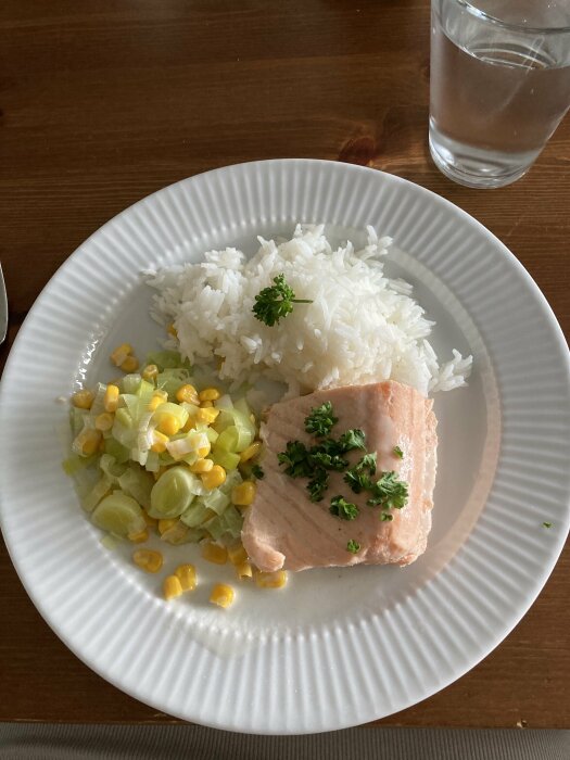En tallrik med ris, lax, majs och purjolök, garnerad med persilja, bredvid ett glas vatten.